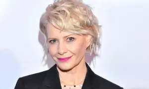 Kobieta z blond włosami w stylowej fryzurze bixie cut, z niebieskimi oczami i różową szminką, w czarnej marynarce.