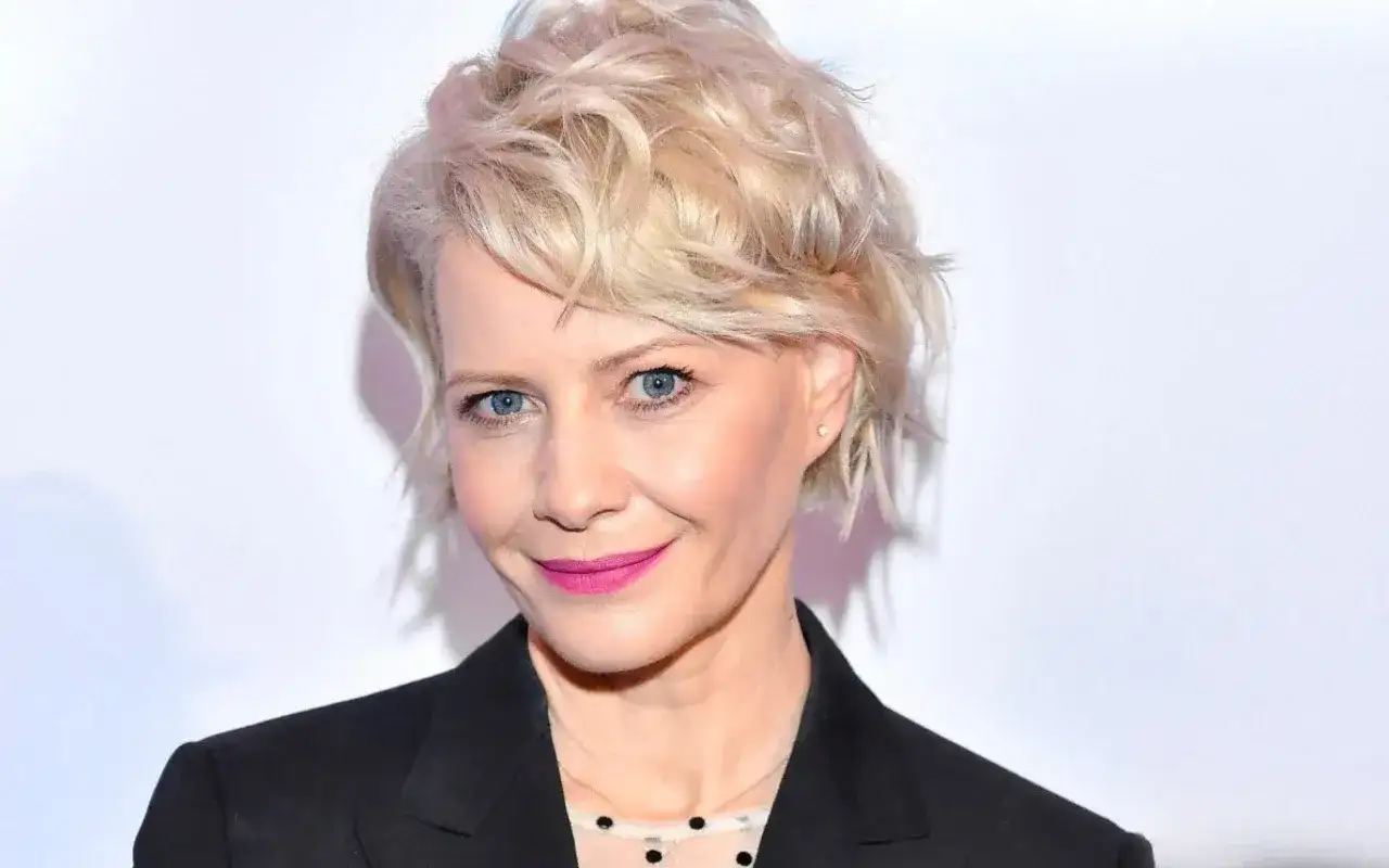 Kobieta z blond włosami w stylowej fryzurze bixie cut, z niebieskimi oczami i różową szminką, w czarnej marynarce.