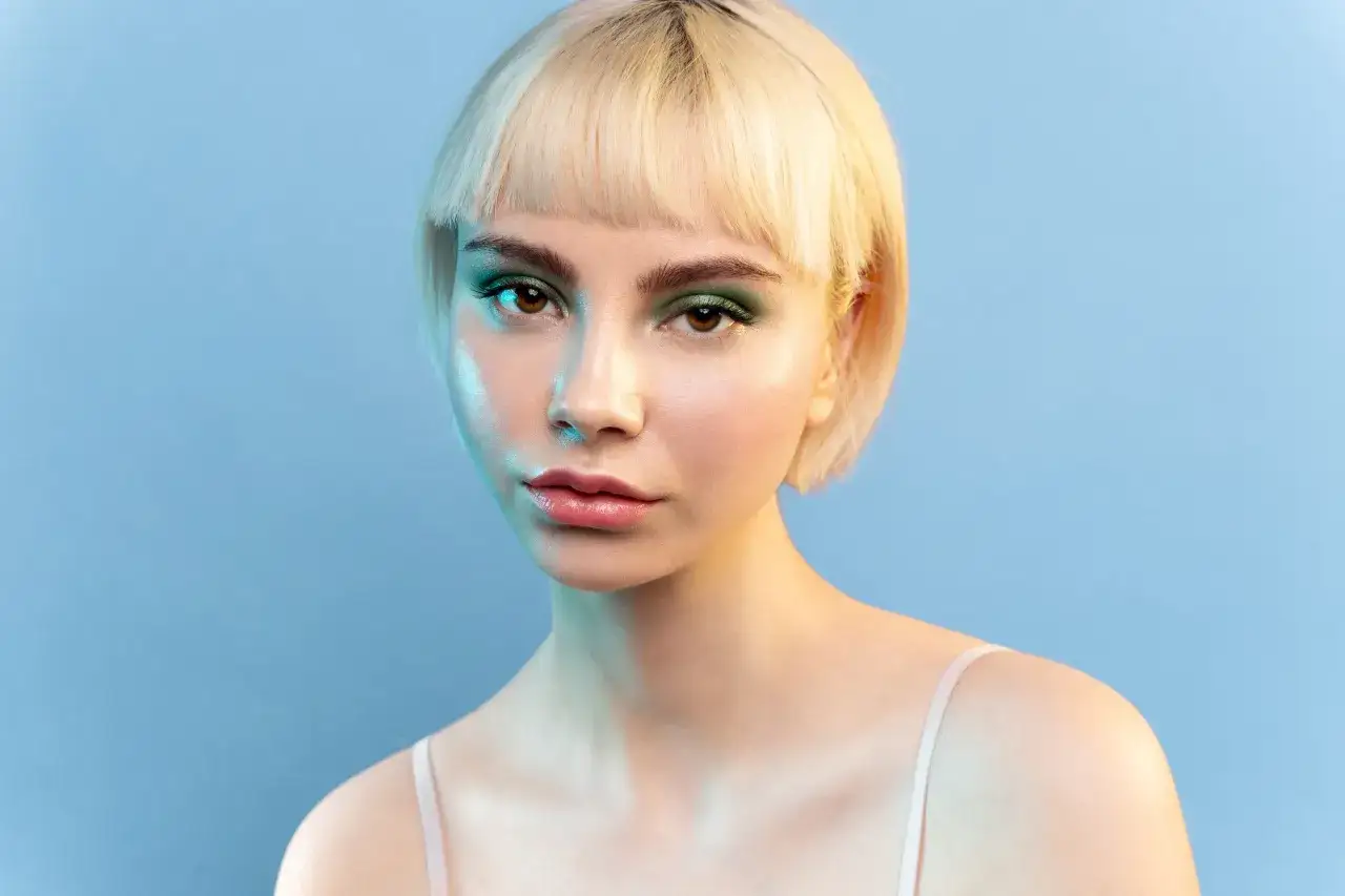 Blondynka z fryzurą na grzybka i zielonym makijażem oczu, oświetlona niebieskim i żółtym światłem.