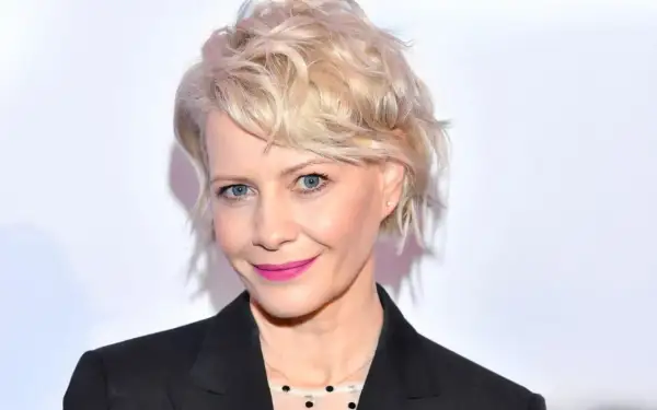 Kobieta z blond włosami w stylowej fryzurze bixie cut, z niebieskimi oczami i różową szminką, w czarnej marynarce.